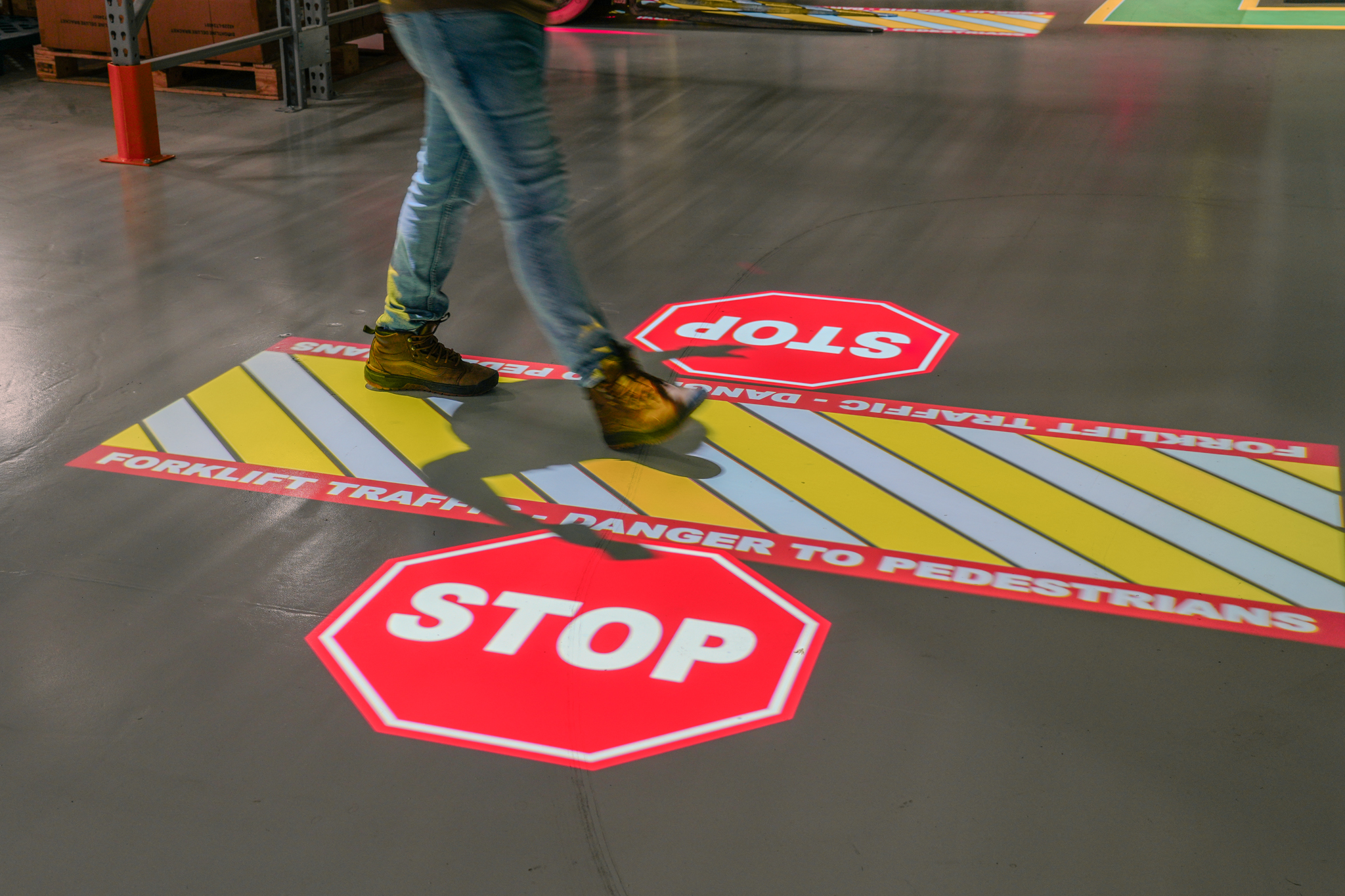 A worker walks across a warehouse floor where projected stop signs and a striped pedestrian warning zone are clearly displayed.
